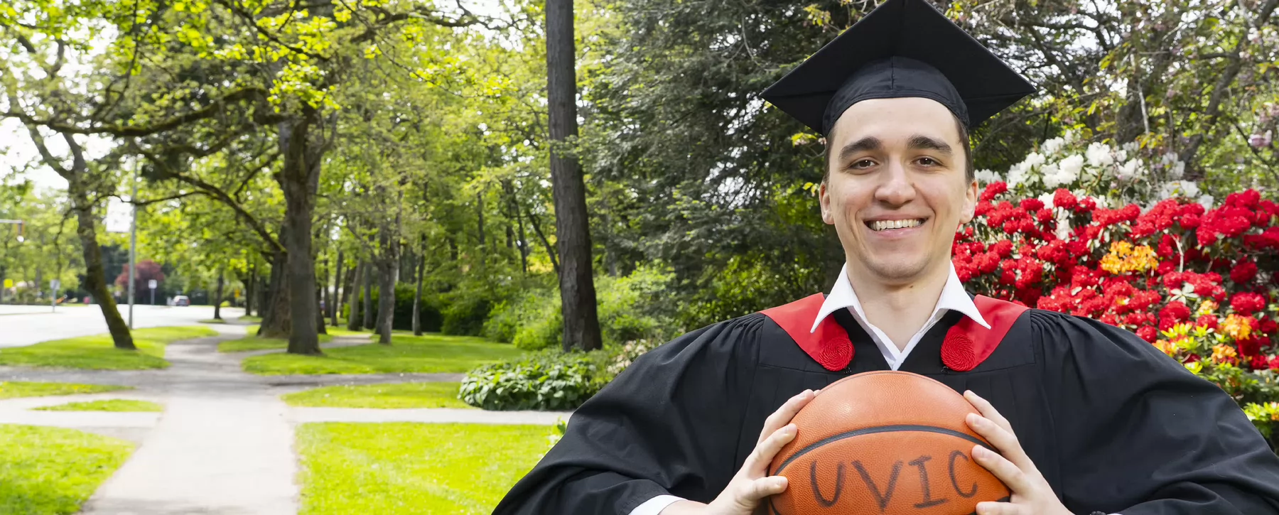 Photo of man with dark hair wearing black graduation robe with red stole holding a basketball out front in both hands