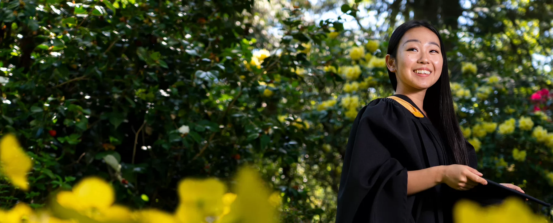 Young asian woman in graduation cap and gown smiling in a park with lush greenery and flowers.