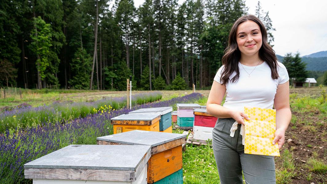 A woman stands in a field lined by trees, next to a group of beehives. She is holding beeswax food wrap.