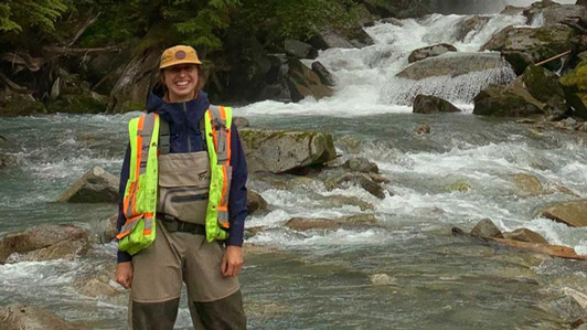 A student wearing a reflective jacket and waders stands in front of a river.