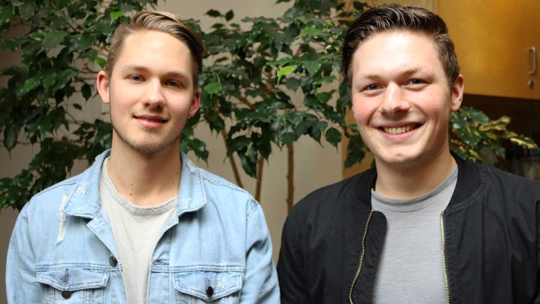 A close-up shot of two men standing in front of a bush. They are both smiling and looking at the camera.