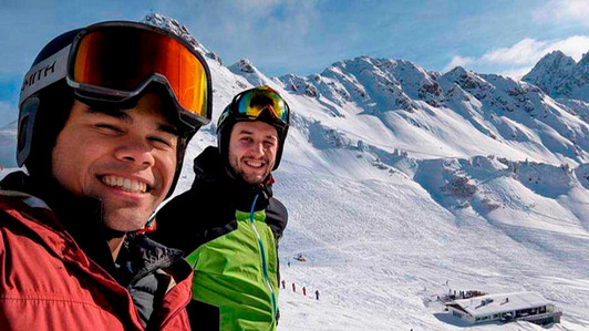 Two students stand on a ski hill wearing goggles and ski suits.