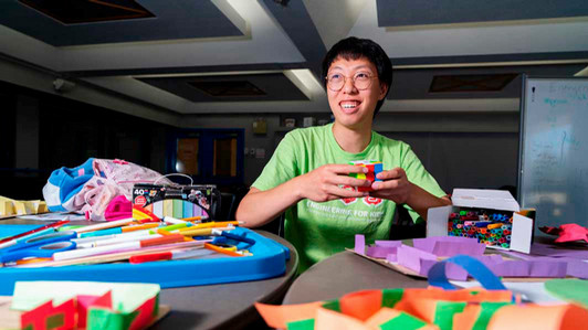 A student holds a Rubik's cube. There are many colourful engineering toys in front of him.
