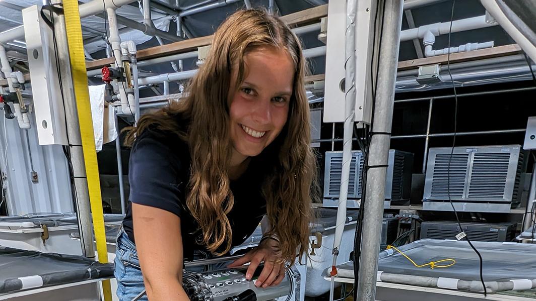 Emily Fricksa is working in a lab, leaning over a table of coral samples, scanning them with a tool and smiling.