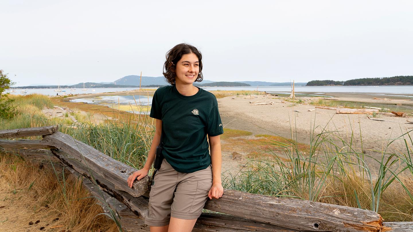 Co-op student Grace Decker is sitting on a wooden fence railing on Sidney Island, wearing a green Parks uniform collared shirt. The beach and the Salish Sea, as well as other small islands, are visible behind Grace.