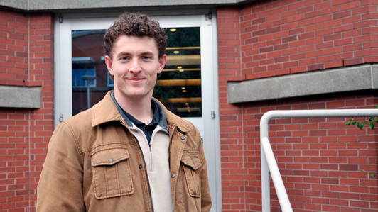 Henry King stands in front of the David Strong Building. The building is made of red brick. Henry wears a light brown jacket. 