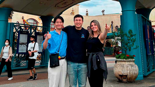 Three people stand together on a street with flowers behind them.