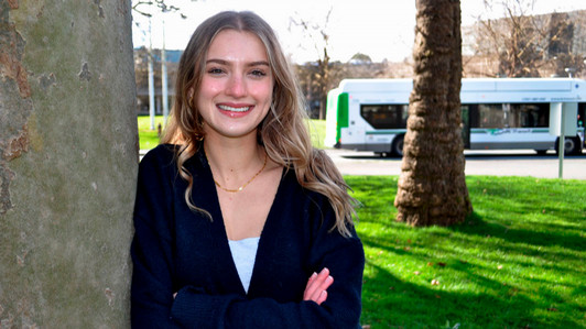 A student stands with her arms folded in front of a tree.