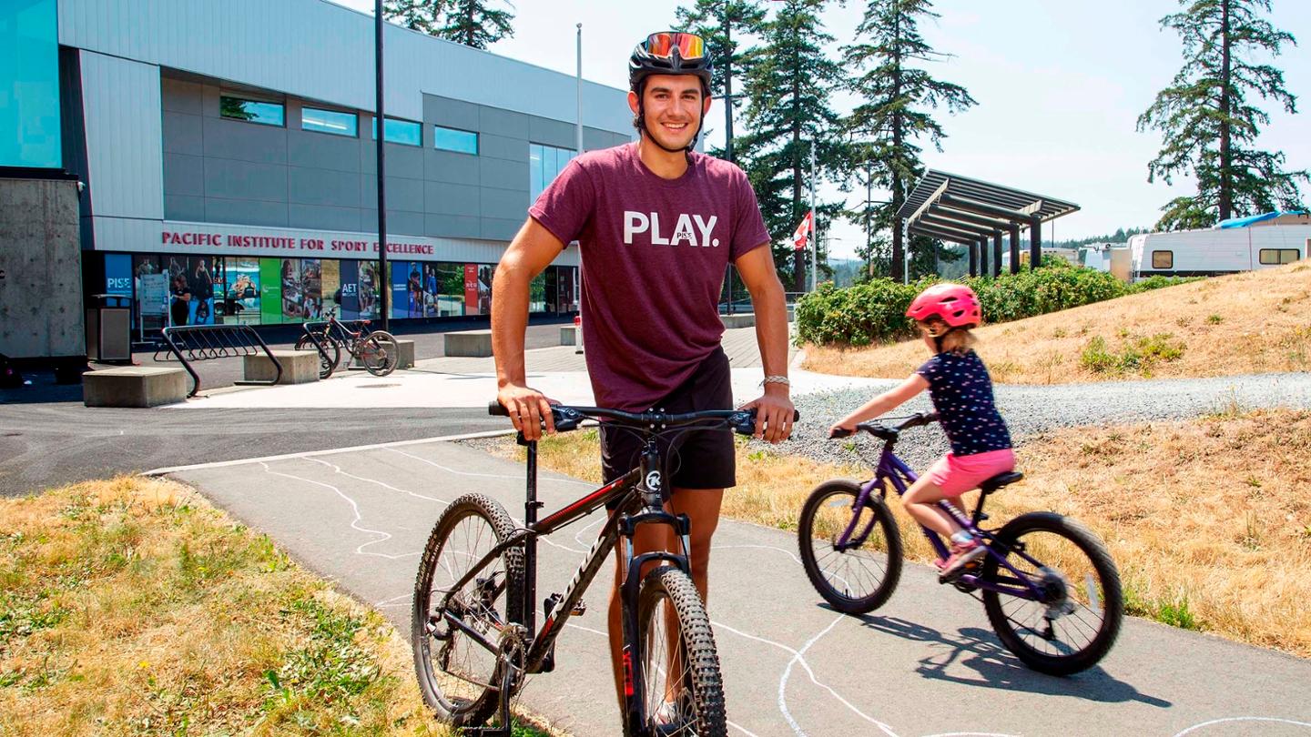A male student stands on a bicycle pathway. He wears a helmet and has a bike at his side. There is a child riding a bike beside him.