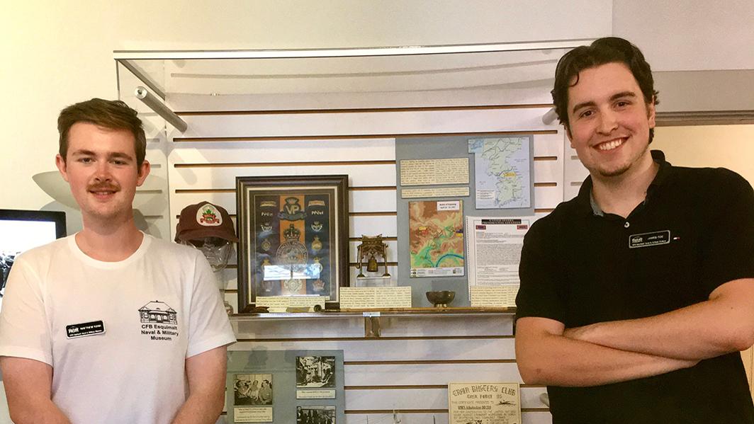 Matthew Kerr (left) and James Coe (right) are standing in front of a curated exhibit on the Korean War, with artifacts in a glass case.