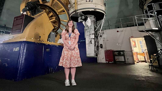 A student wearing a dress stands in front of equipment in an observatory