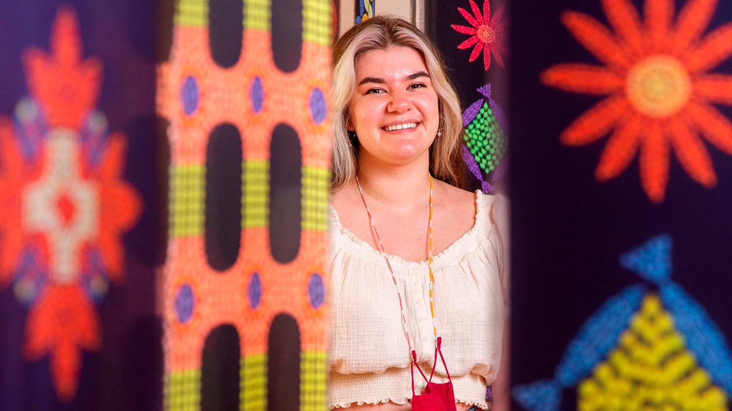 A student stands in between several colourful beaded art pieces that are hanging from the ceiliing.