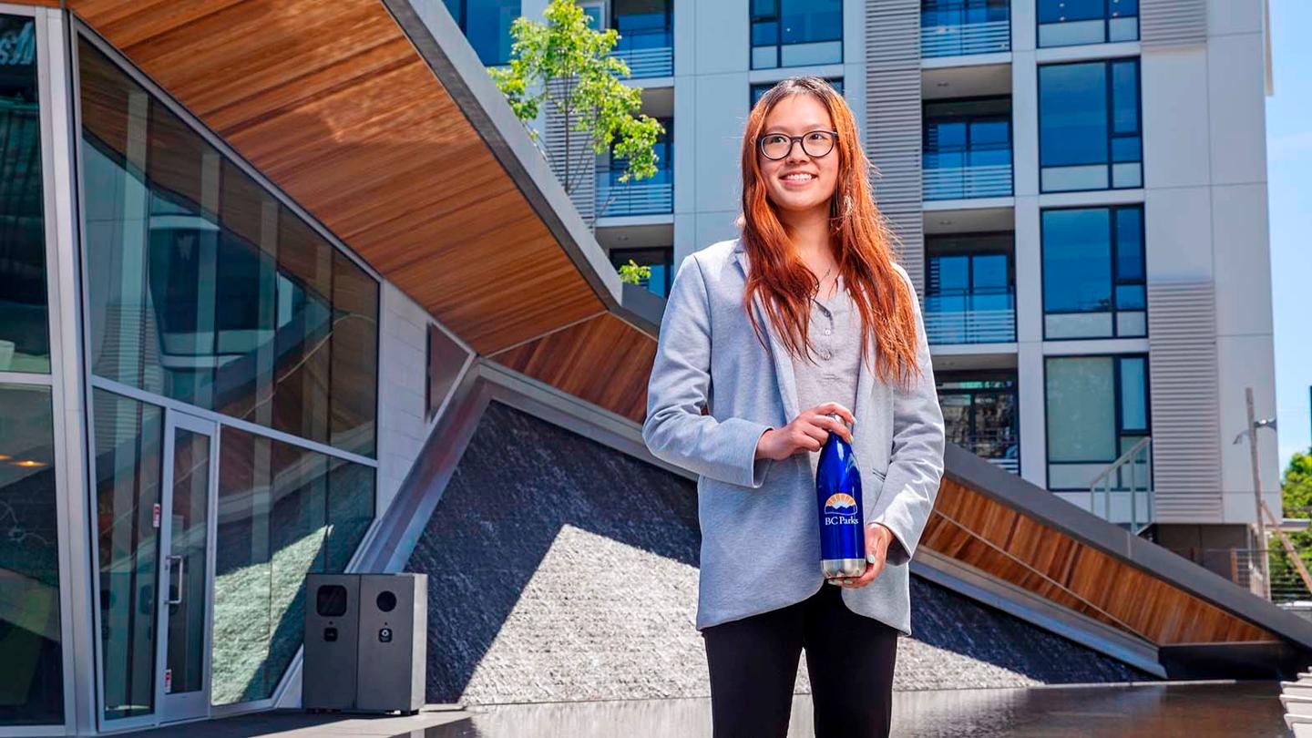 A student holding a water bottle with the BC Parks logo stands in front of a wood and glass building.