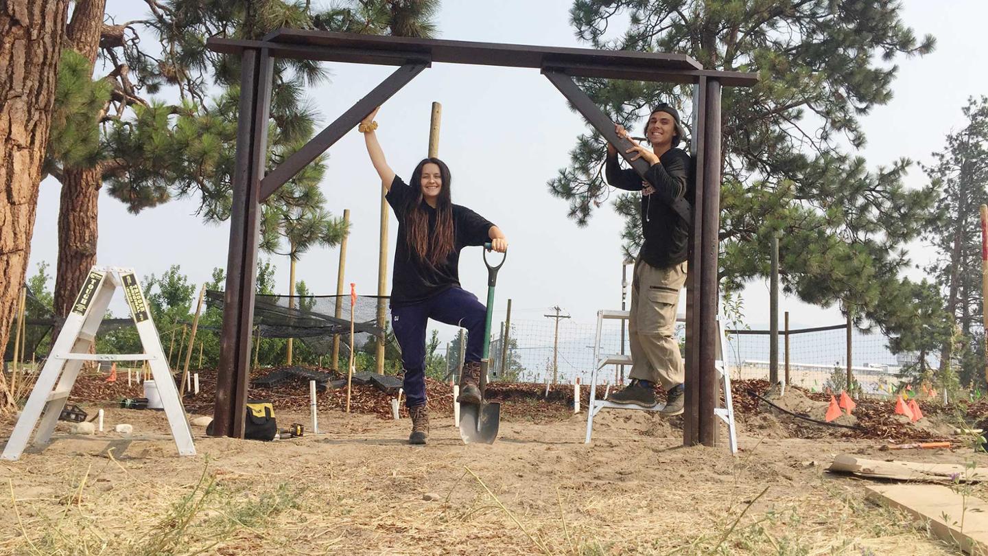 Two students stand at the entrance to an Indigenous garden. There is a wooden arbour behind them. These students are Dana Johnson and Kyle Clarke.