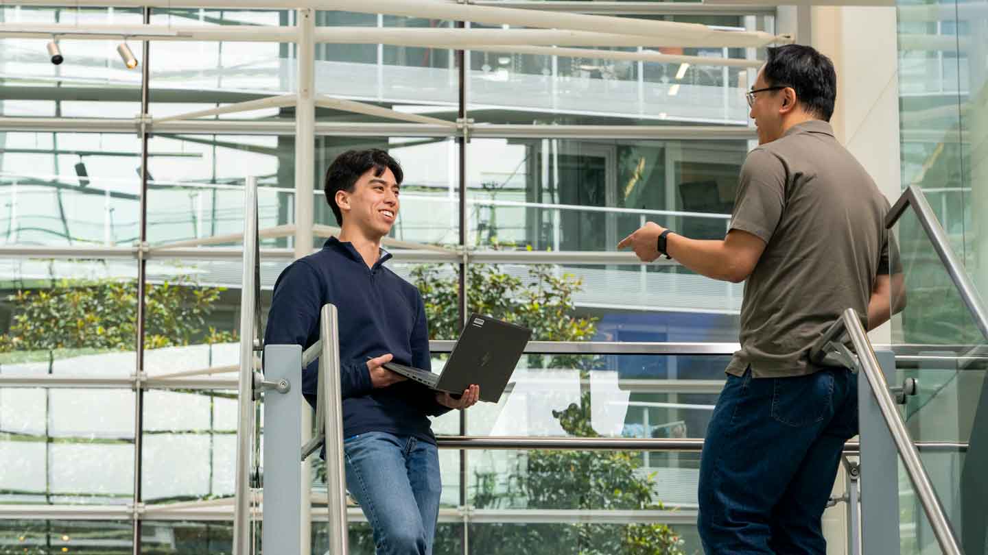 A student and his employer stand at the top of a staircase in an atrium with many windows. The student is holding a laptop.