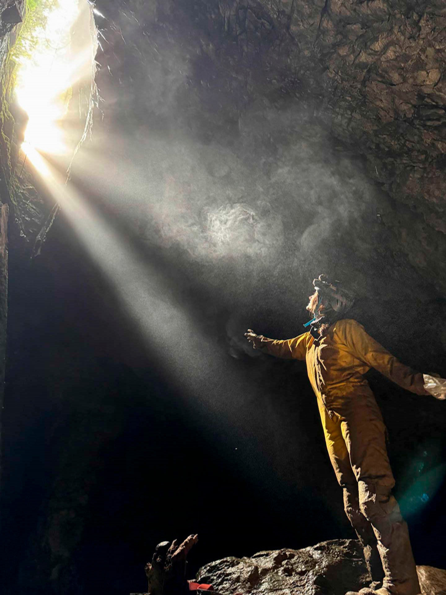 A student wearing climbing gear stands in a cave with light coming from above.