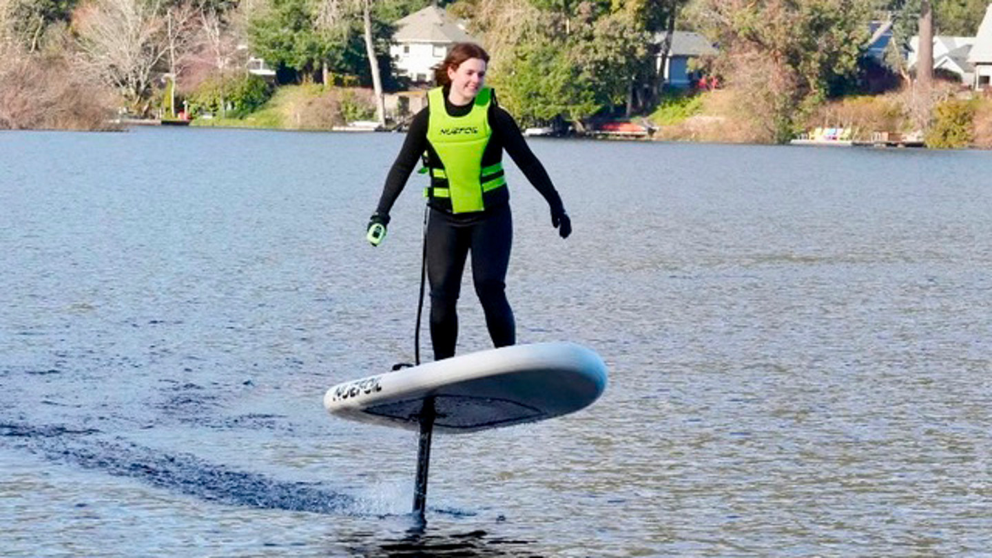 A woman wearing a bright green life jacket stands on a e-foil board on a body of water