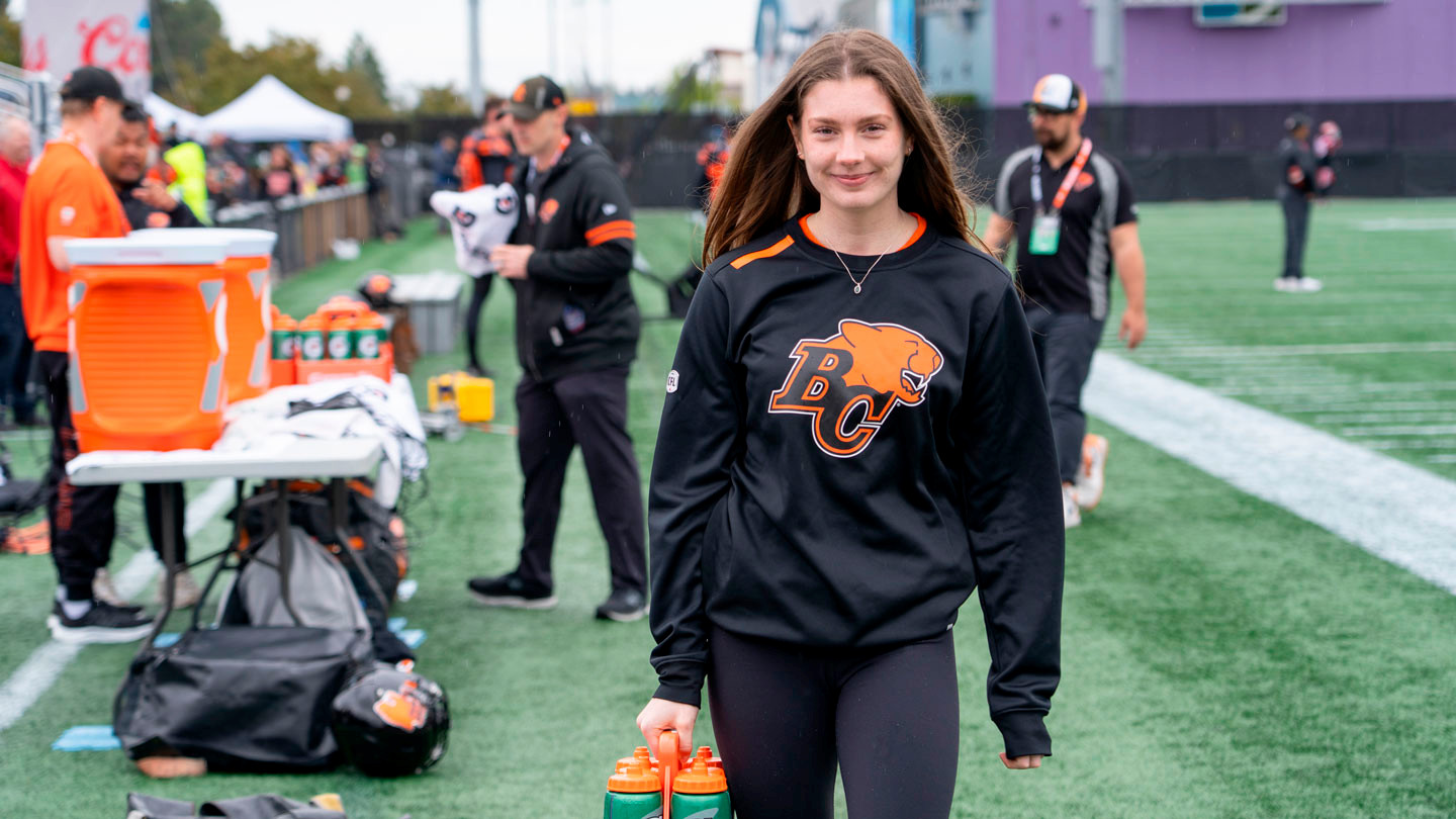 A female student with long brown hair wearing a BC Lions jersey and holding gatorade water bottles walks across a football field during a BC Lions practice.