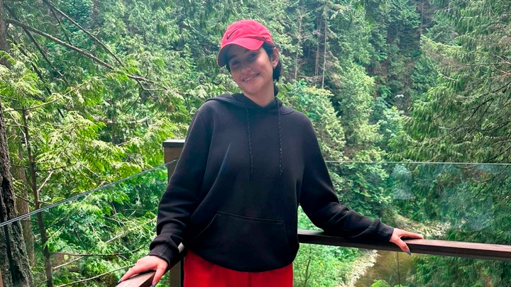 A student wearing a red hat stands in from of a forest.