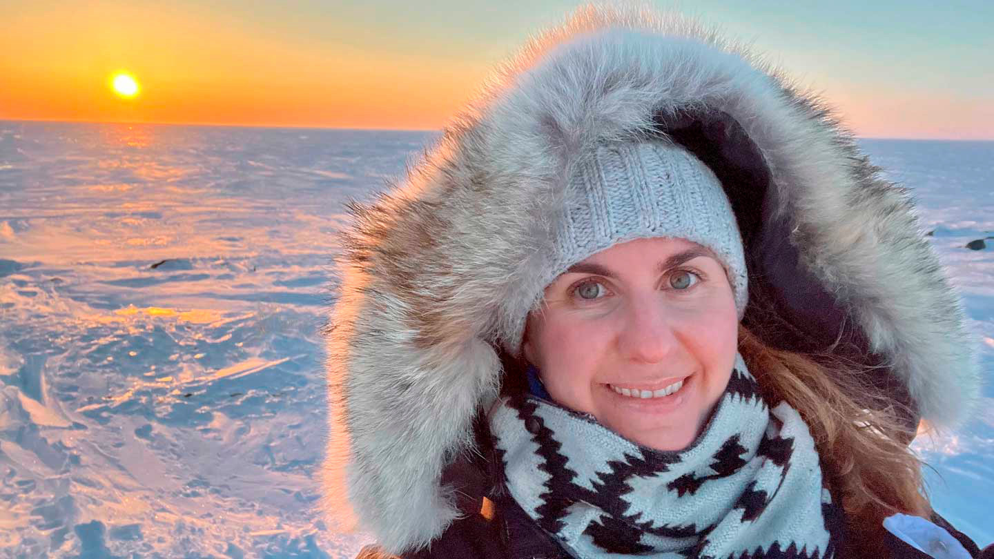 A woman wearing a fur-lined parka stands in front of a snowy landscape.