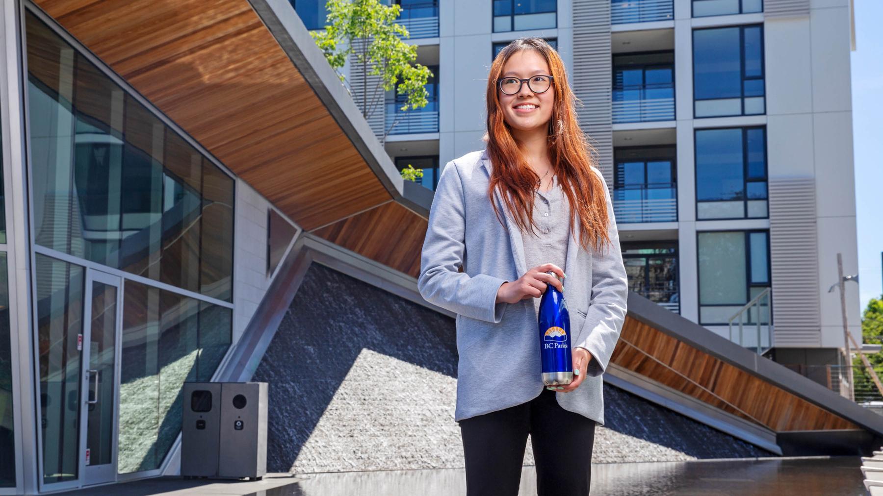 A student is standing on the UVic campus, holding a BC Parks branded water bottle. She is smiling at the camera.
