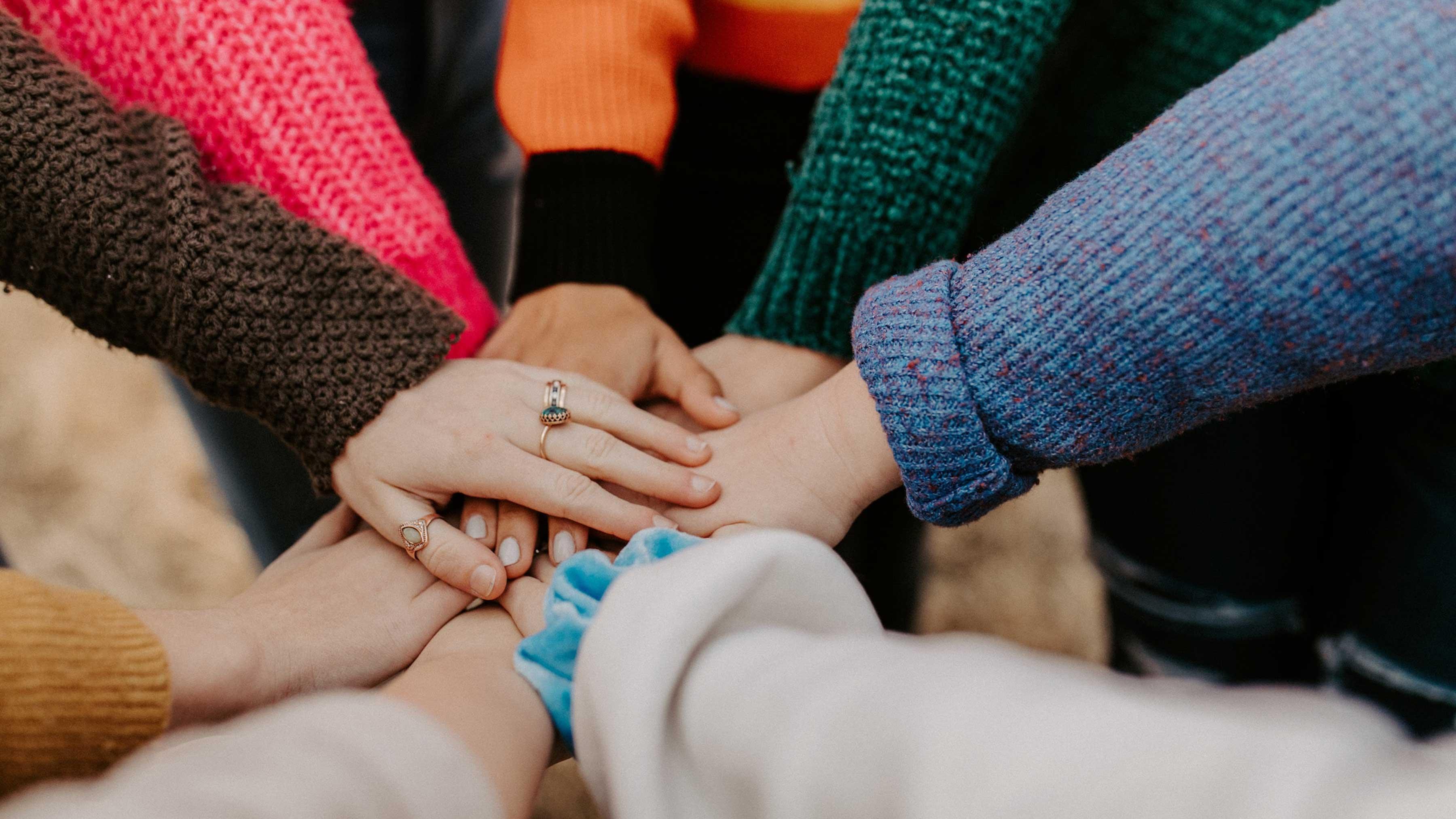 A close-up of many students' hands coming together in a circle. Each student is wearing a different colour of sweater.