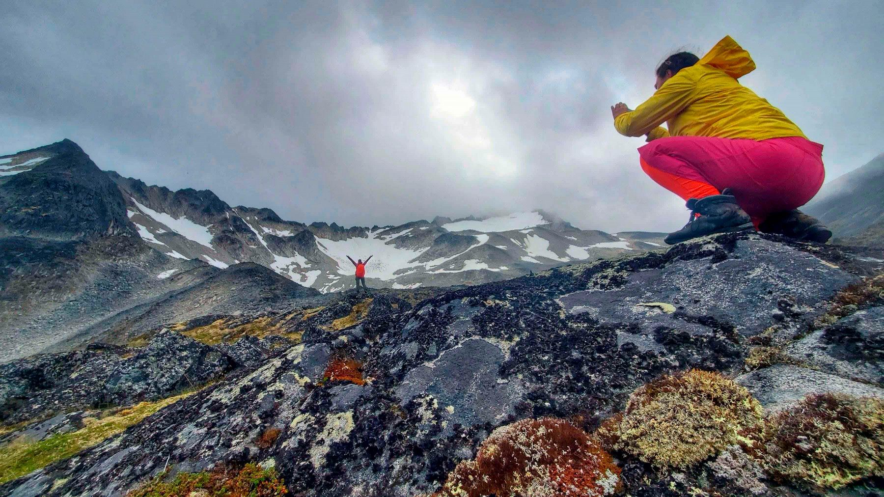 A student wearing bright hiking gear takes a photo of another student on the top of a mountain.