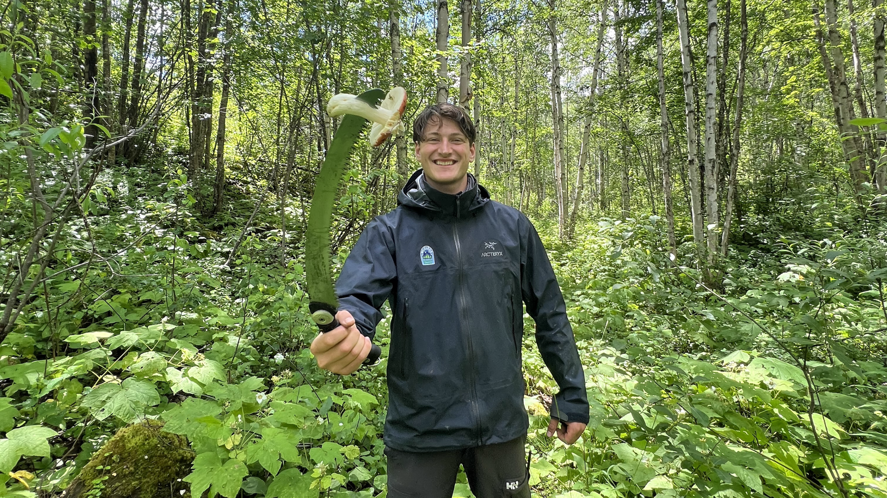Karl Hare, a co-op student, is standing very happily in the forest, dressed in a waterproof black jacket and black trousers. He is holding up a large serrated knife, on which is impaled a giant white mushroom. He is grinning widely.