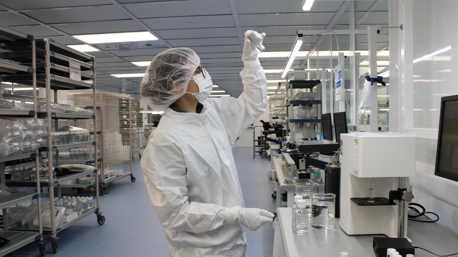 Mariana Latta Suazo, a UVic co-op student, is working in a lab, wearing a protective lab coat, hairnet, gloves, face mask and glasses. Mariana is holding up a small vial to examine its contents.