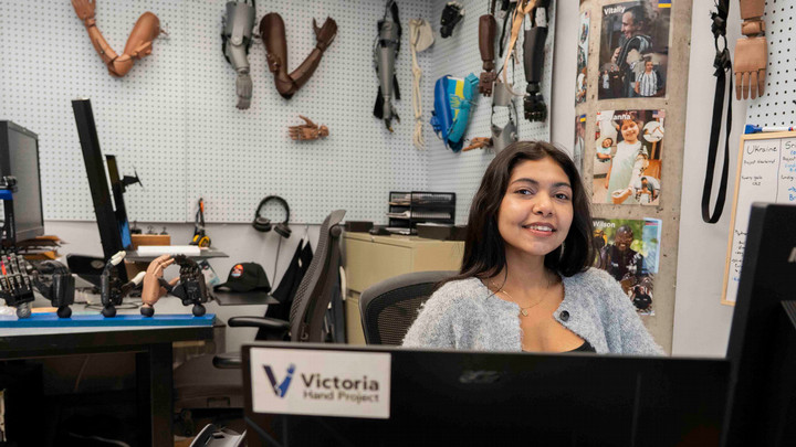 A student sits behind a desk. There are prosthetic hands on the wall behind her.