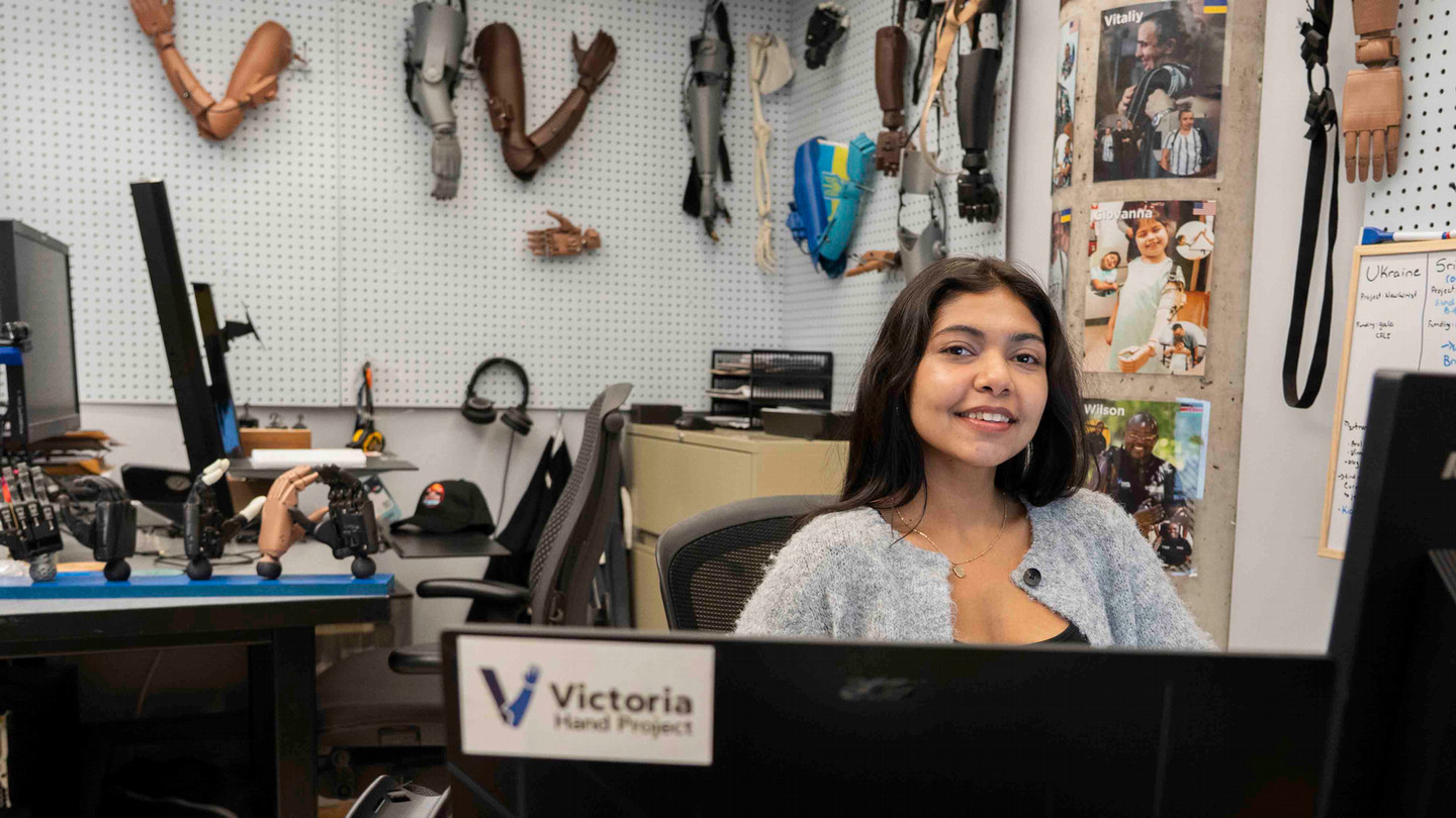 A student sits behind a desk. There are prosthetic hands on the wall behind her.