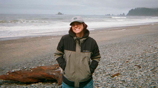 A student wearing a jacket and hat stands on a beach on a cloudy day