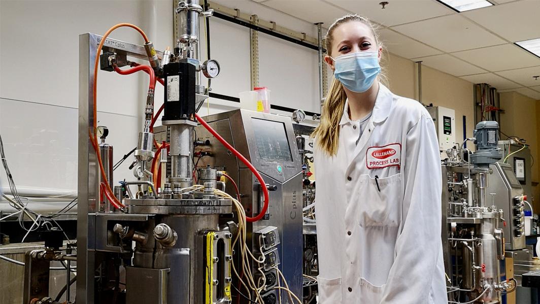 A woman wearing a Lallemand lab coat and a mask stands in a room full of yeast processing equipment.
