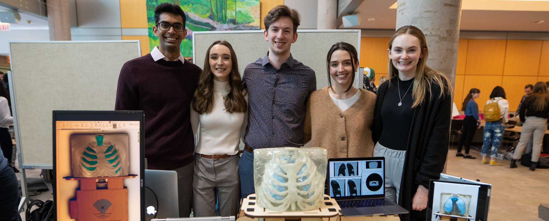 Two male and three female students stand smiling linked together behind a table displaying various electronic and physical models of a ribcage and lungs.  Other displays in the ELW lobby can be seen in the background.