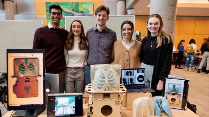 Five students stand in a lobby behind a table, where their engineering capstone project sits.