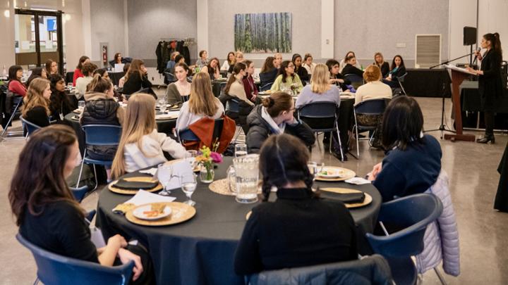 Women seated around tables in a dining hall listen to a person speaking at a podium.