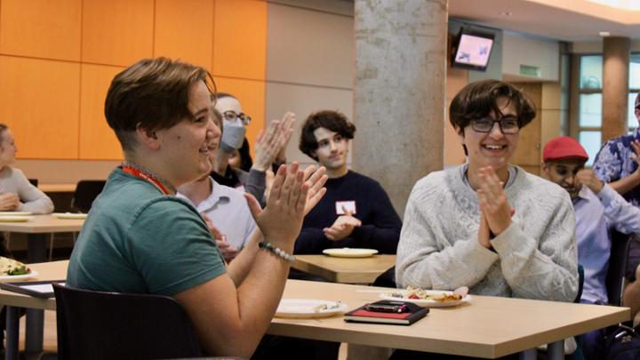 Several students are showing sitting at tables and applauding.