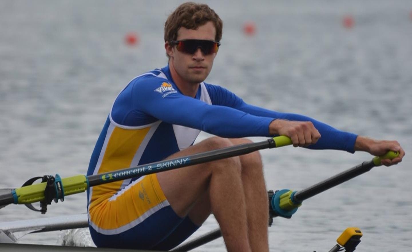 Giancarlo wearing sunglasses sits in his rowing shell ready to stroke on a lake with course markers