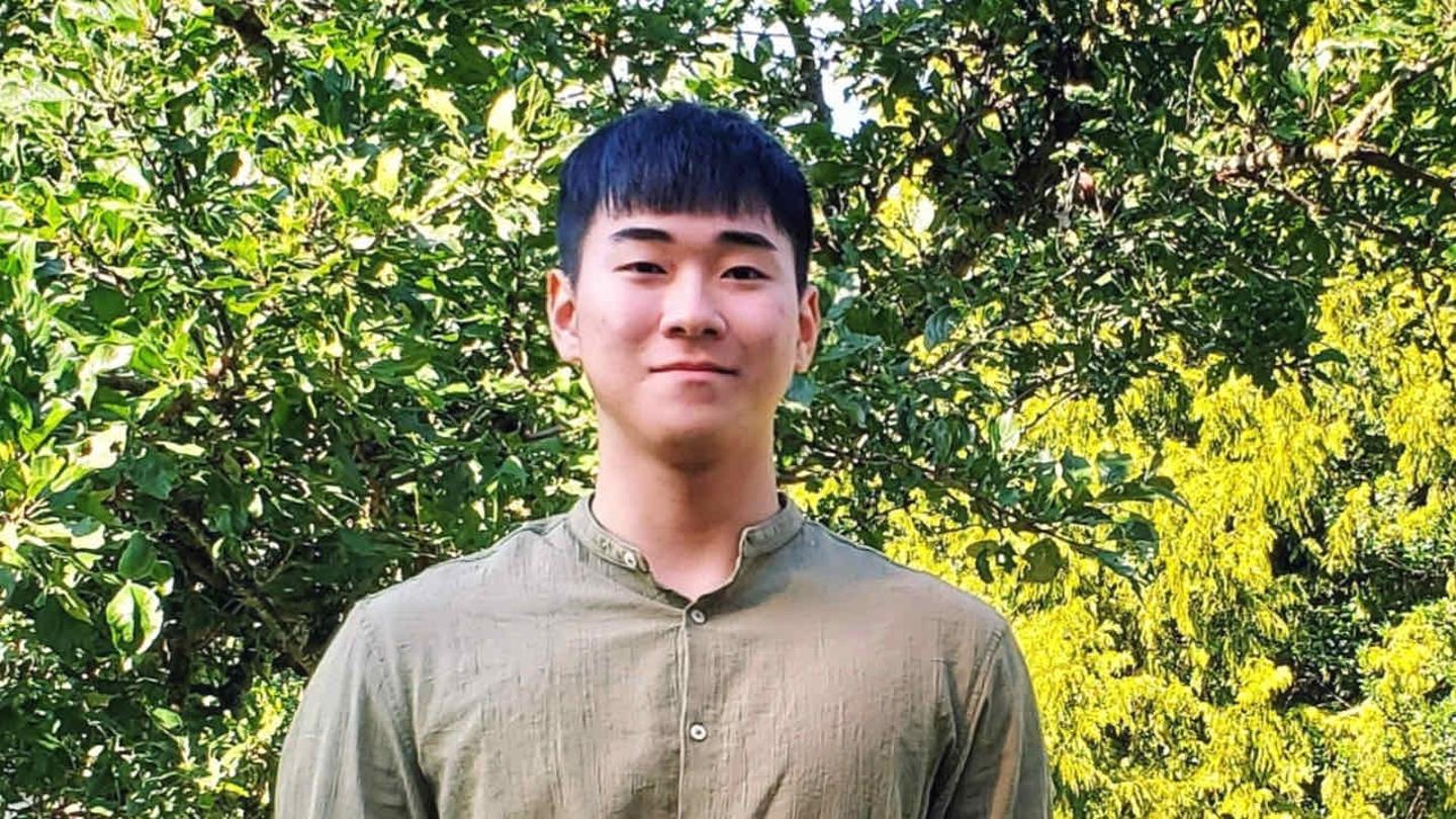 Hanuel Jang stands outdoors, with trees and bushes behind him.