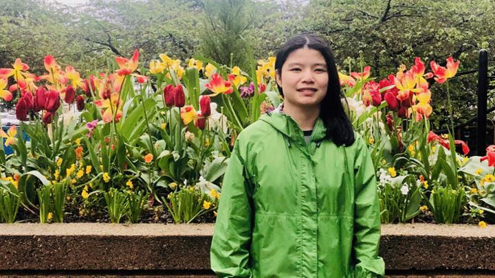 A young woman stands outdoors in front of a colourful bed of flowers.