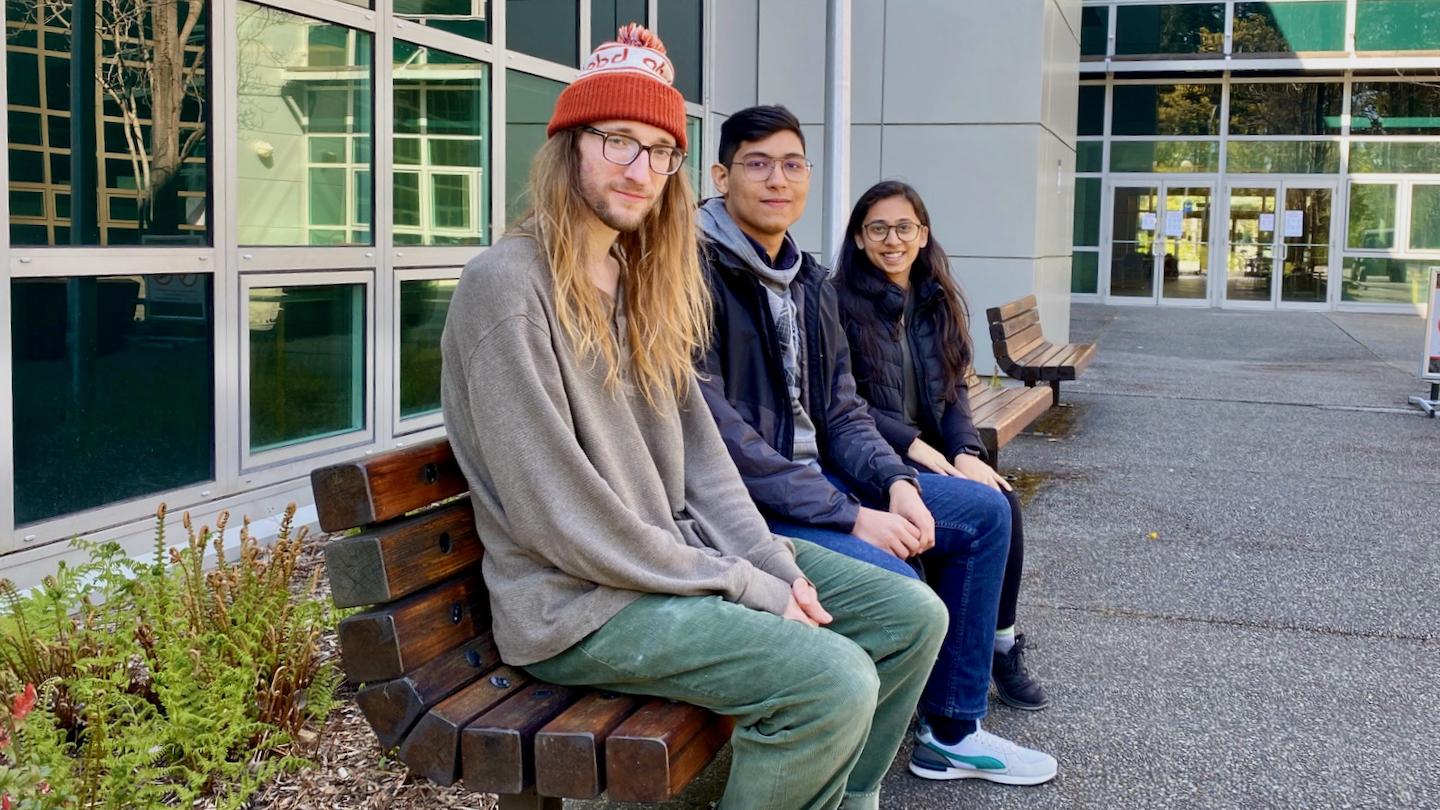 Three students sit on a bench outside a building on campus.