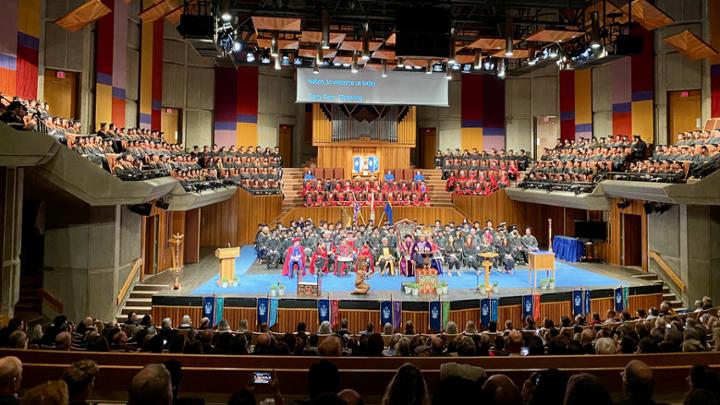 A stage and the seats behind it are filled with hundreds of students and officials dressed in university regalia.