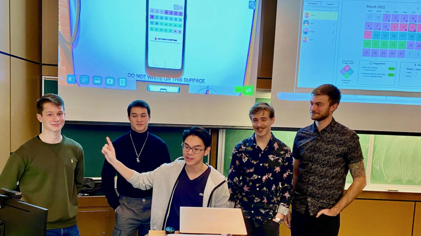 Five students present their startup at the front of a classroom, standing behind a podium with a laptop. Two large overhead projector screens in the background show a cellphone and a digital calendar.