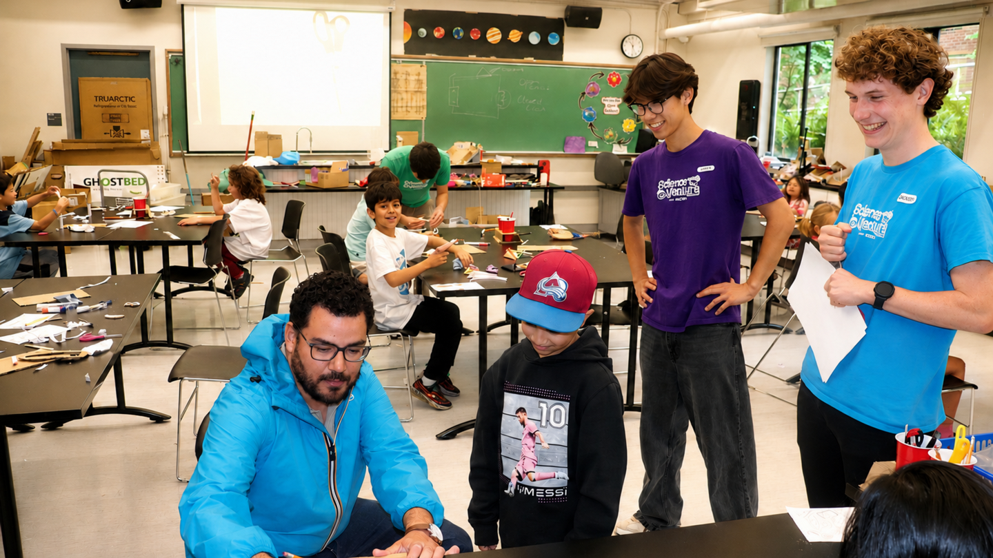 MP Will Greaves in a classroom signing an autograph with children at tables in the background.