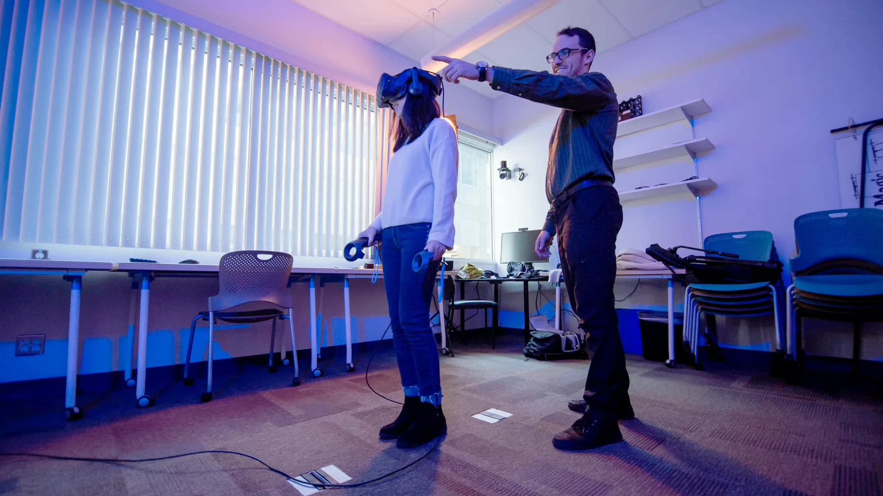 A woman wearing a VR headset with a man standing beside her pointing at something.