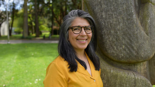 An head-and-shoulders portrait of Dr. Claudia Diaz, pictured outdoors on campus. She is wearing a yellow blouse and tortise shell glasses, facing the camera, and smiling. 