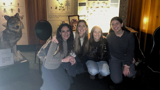 Four smiling women crouch together in a dimly lit room, with a taxidermy wolf and nature displays behind them. The atmosphere is warm and friendly.