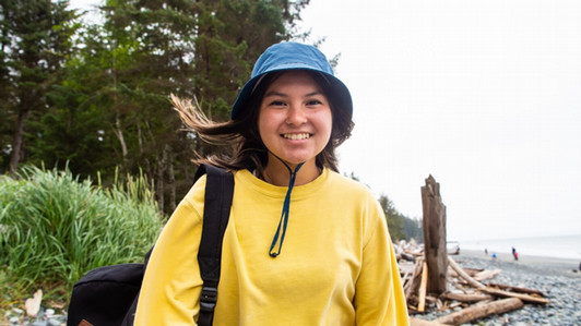 A graduate student pictured doing research at the beach. They are facing the camera and smiling, wearing a yellow sweater and a blue bucket hat..