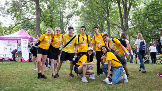 A group of students in yellow t-shirts smiling toward the camera. The location is outdoors on campus during the summer season.