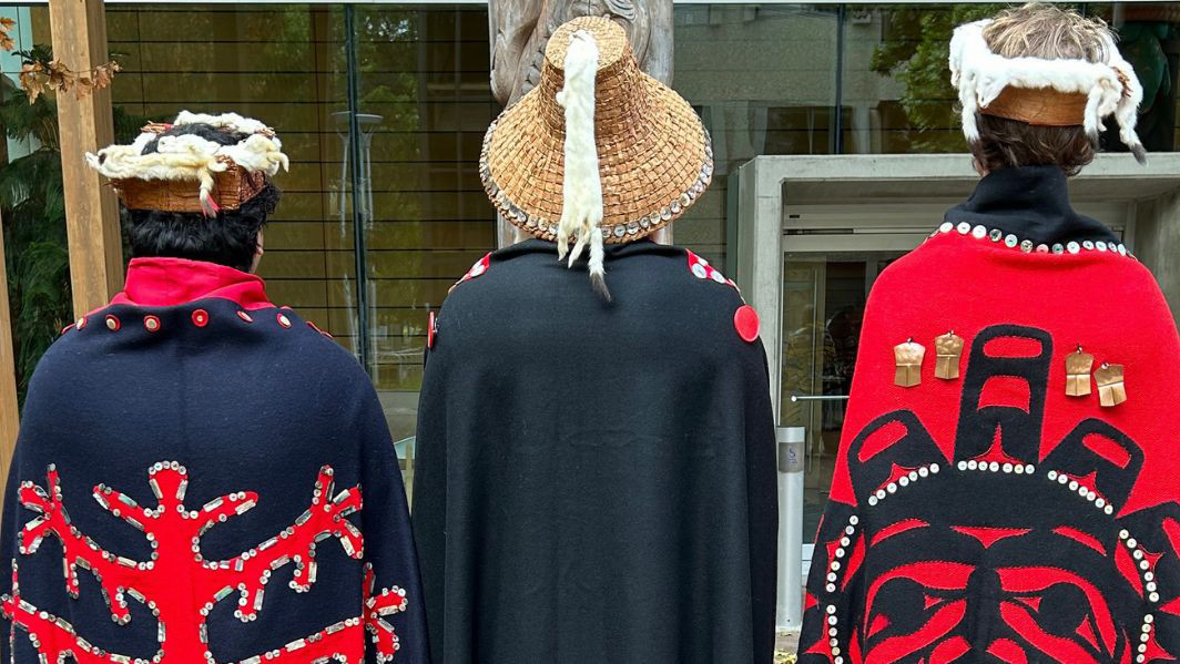 Three people in Indigenous dress are pictured outside First Peoples House on campus. They are facing away from the camera with the focus of the image on the designs of the back of their clothing. Each outfit is different.